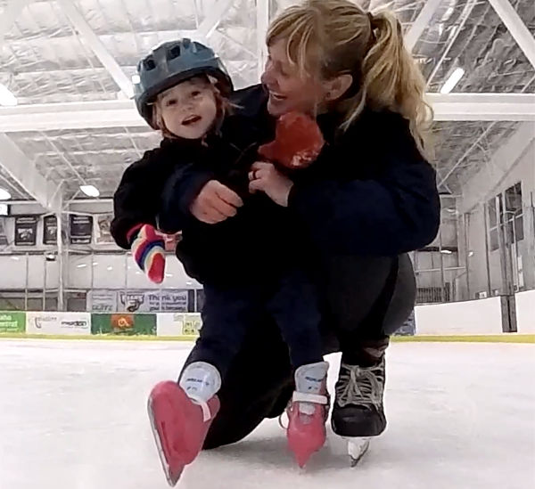 Rink manager Laura Wold kneeling on the ice, holding up a laughing young skater in a helmet and pink skates on her first real strides.